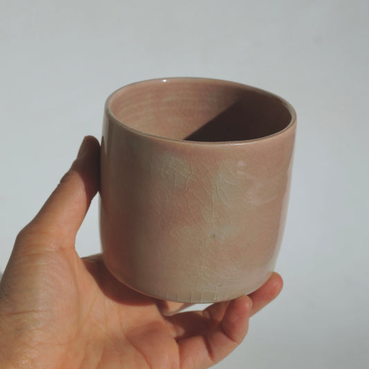 A hand holding a small cylindrical ceramic cup with a dusty pink glaze and subtle crackled texture against a plain light gray background.
